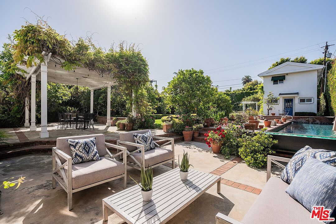 This inviting backyard space features a concrete patio area furnished with comfortable outdoor seating and a slatted coffee table. A white pergola covered in lush greenery provides shade for a dining area, while a swimming pool sits adjacent to a small, white two-story structure. The scene is bathed in bright, natural sunlight, creating a serene and well-maintained outdoor living environment.