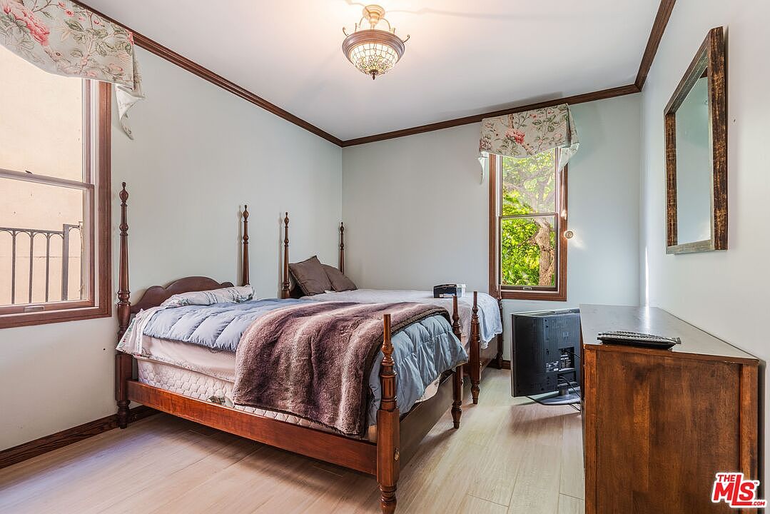 This guest bedroom features two wooden four-poster beds with blue bedding and dark brown throws, set against light-colored walls. The room is illuminated by a decorative ceiling light fixture and natural light from two windows adorned with floral valances. A wooden dresser sits to the right, topped with a television, while a mirror hangs on the wall above it.