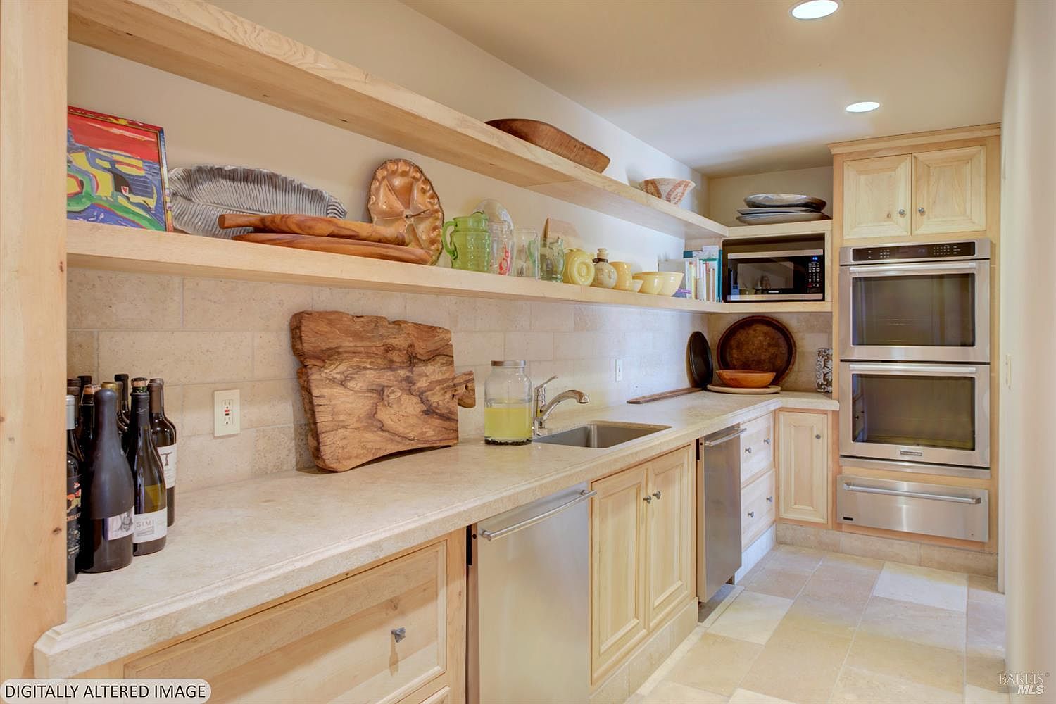 This kitchen features a warm, rustic aesthetic with light wood cabinetry, natural stone countertops, and open shelving displaying decorative pottery and glassware. The space is equipped with a double wall oven, a built-in microwave, and a stainless steel sink, all set against a textured stone backsplash. The perspective is a straight-on eye-level shot that highlights the functional layout and the cozy, inviting atmosphere of the culinary space.