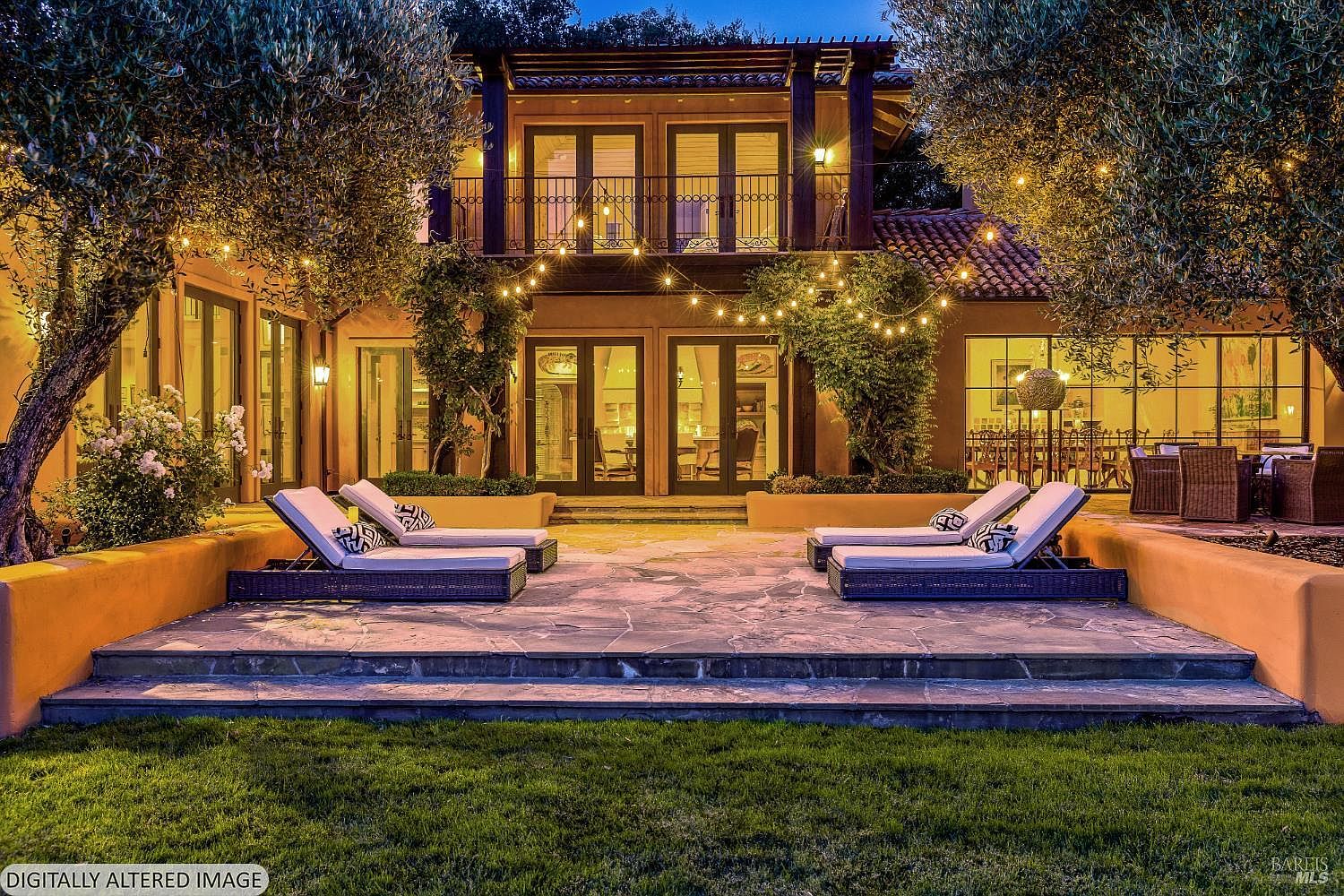 This elegant rear exterior view showcases a Mediterranean-style home illuminated by warm string lights at dusk. The scene features a stone patio with four lounge chairs, framed by manicured olive trees and a low, warm-toned retaining wall. The perspective is centered and eye-level, highlighting the symmetrical architecture, balcony, and inviting indoor-outdoor flow.