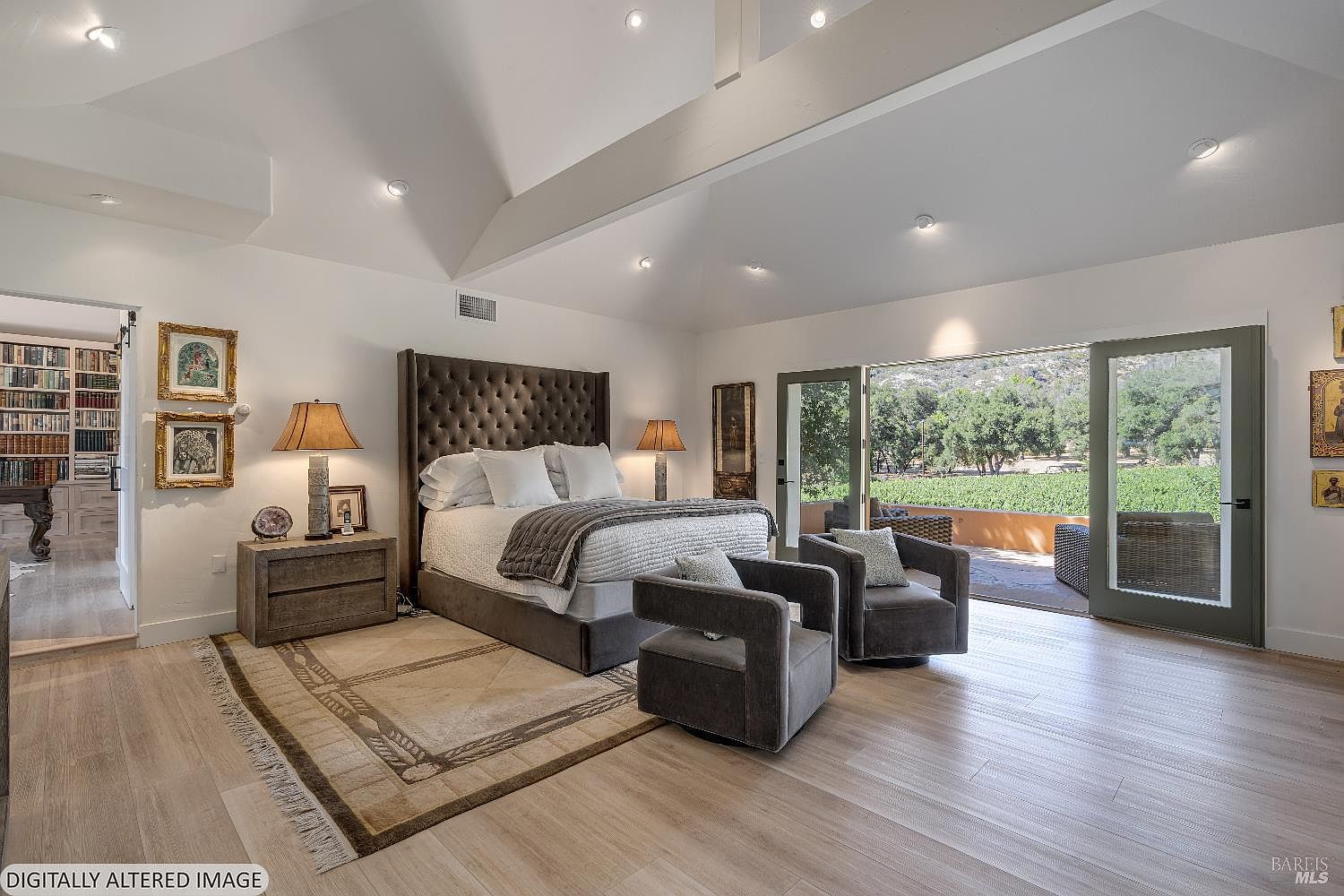 This spacious primary bedroom features a high vaulted ceiling with exposed beams and light-toned hardwood flooring, creating an airy and luxurious atmosphere. A large, tufted velvet headboard serves as the focal point, complemented by two modern armchairs and a view through wide glass doors that open onto a scenic outdoor patio. The room also includes a dedicated library or office space visible through an adjacent doorway, enhancing the sophisticated and tranquil feel of the suite.