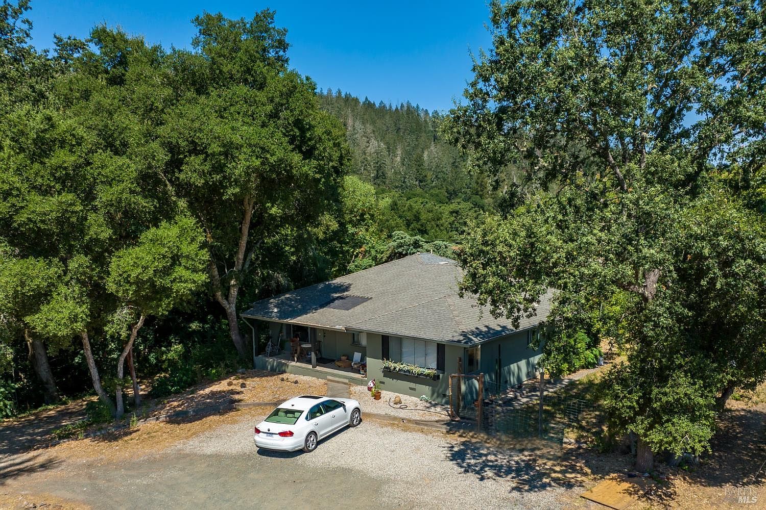 This aerial perspective captures a single-story home nestled in a lush, wooded landscape with a backdrop of rolling, tree-covered hills. The house features a simple, functional design with a covered front porch and a gravel driveway where a white sedan is parked. The surrounding mature trees and natural terrain create a secluded, peaceful atmosphere, highlighting the property's integration with its rural environment.