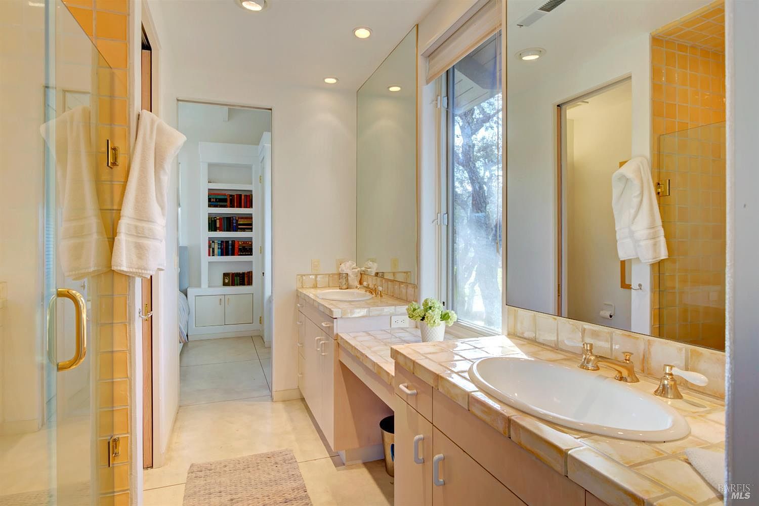 This bright, dual-vanity bathroom features light-toned cabinetry topped with warm, earth-toned tiled countertops and integrated sinks. A large mirror spans the wall above the vanity, reflecting the natural light from the adjacent window, while a glass-enclosed shower is visible on the left. The space leads into a hallway with built-in shelving, creating a functional and airy atmosphere.