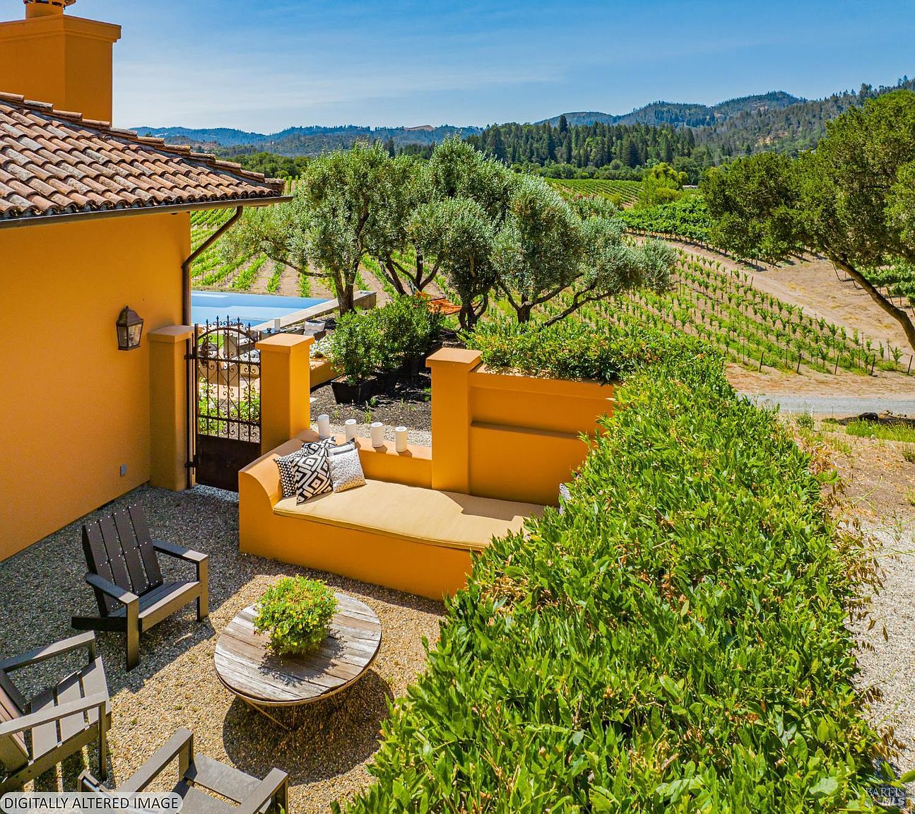 This inviting outdoor patio area features a built-in yellow stucco bench with plush seating, set against a backdrop of lush greenery and a sprawling vineyard. The space includes a rustic wooden coffee table and dark-toned Adirondack chairs, all situated on a gravel terrace that overlooks the scenic landscape. The warm, Mediterranean-inspired architecture and panoramic views create a serene and luxurious atmosphere perfect for relaxation.