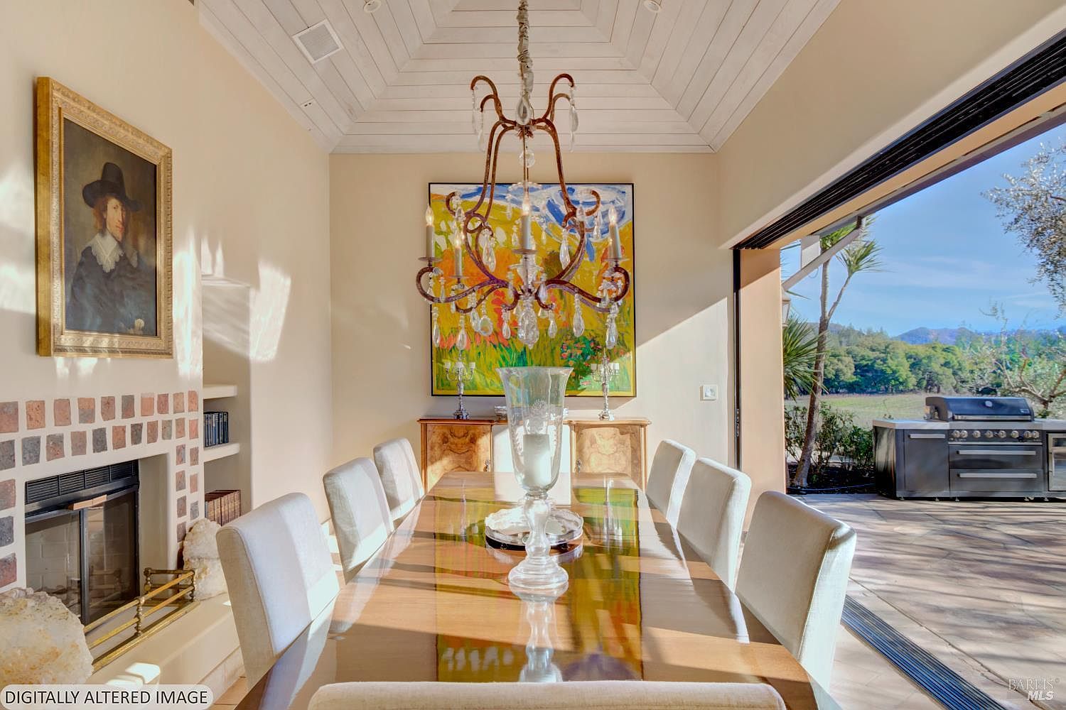 This elegant dining room features a long, polished wooden table surrounded by upholstered chairs, set beneath a grand, ornate chandelier. The space is anchored by a vibrant abstract painting on the back wall and a classic portrait to the left, while a large sliding glass door opens to an outdoor patio with a grill and scenic landscape views. The room exudes a sophisticated, airy atmosphere, blending traditional art with modern indoor-outdoor living.
