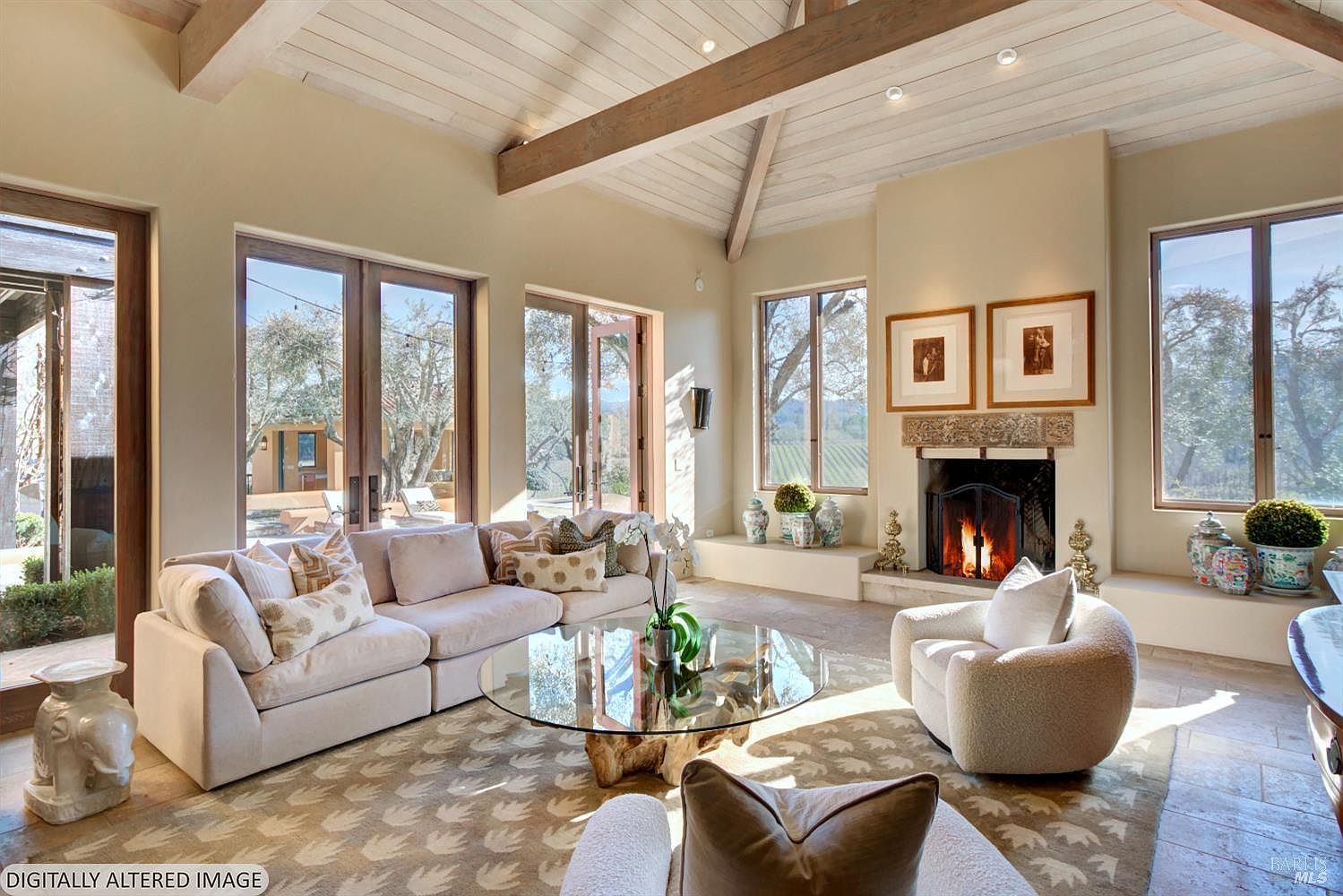 This elegant living room features a high vaulted ceiling with exposed wooden beams and a cozy fireplace as the central focal point. The space is furnished with a plush, light-colored sectional sofa and a modern armchair, all arranged around a unique glass-topped coffee table with a natural wood base. Large windows and glass doors provide abundant natural light and scenic views of the surrounding landscape, creating a warm and inviting atmosphere.