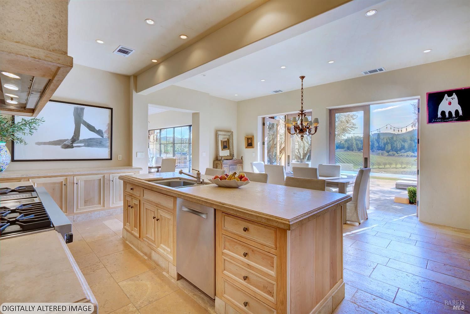 This spacious, light-filled kitchen features a large central island with a stainless steel dishwasher and a sink, complemented by light wood cabinetry and stone countertops. The open-concept design flows seamlessly into a dining area with a chandelier, which opens through large sliding glass doors to a scenic outdoor patio and vineyard view. The aesthetic is warm and inviting, blending traditional cabinetry with a bright, airy atmosphere perfect for entertaining.