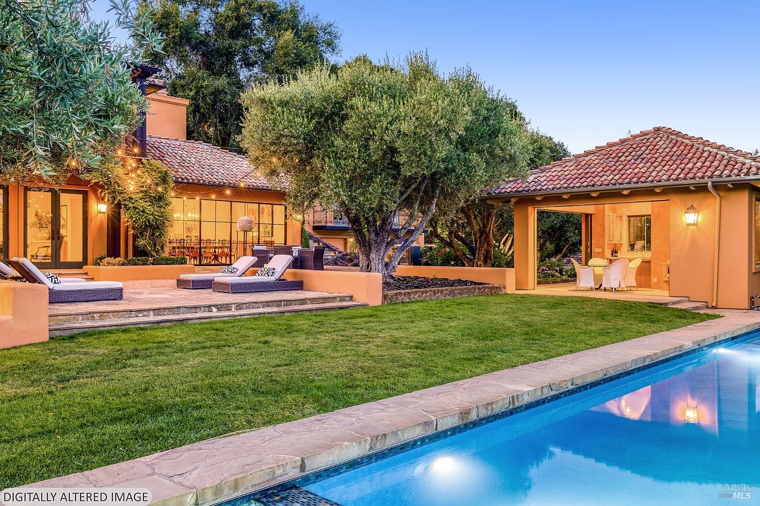 This stunning Mediterranean-style backyard features a sparkling blue swimming pool in the foreground, leading to a spacious stone patio with lounge chairs. A large, mature olive tree serves as a central focal point, flanked by a main residence with expansive glass doors and a separate covered outdoor living area. The warm, golden-hued stucco walls and terracotta roof tiles create a luxurious, inviting atmosphere perfect for outdoor entertaining.