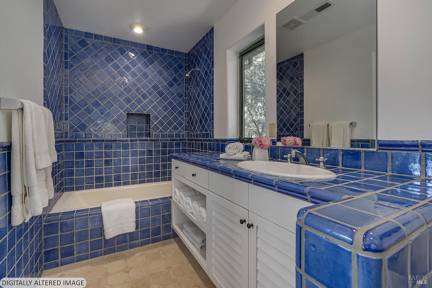 This vibrant bathroom features striking cobalt blue square tiles covering the walls, bathtub surround, and vanity countertop, creating a bold, Mediterranean-inspired aesthetic. The space includes a white vanity with open shelving for towels, a clean white sink, and a large mirror reflecting the room's unique character. The perspective is a wide-angle shot from the doorway, capturing the contrast between the deep blue tile work and the crisp white cabinetry and fixtures.