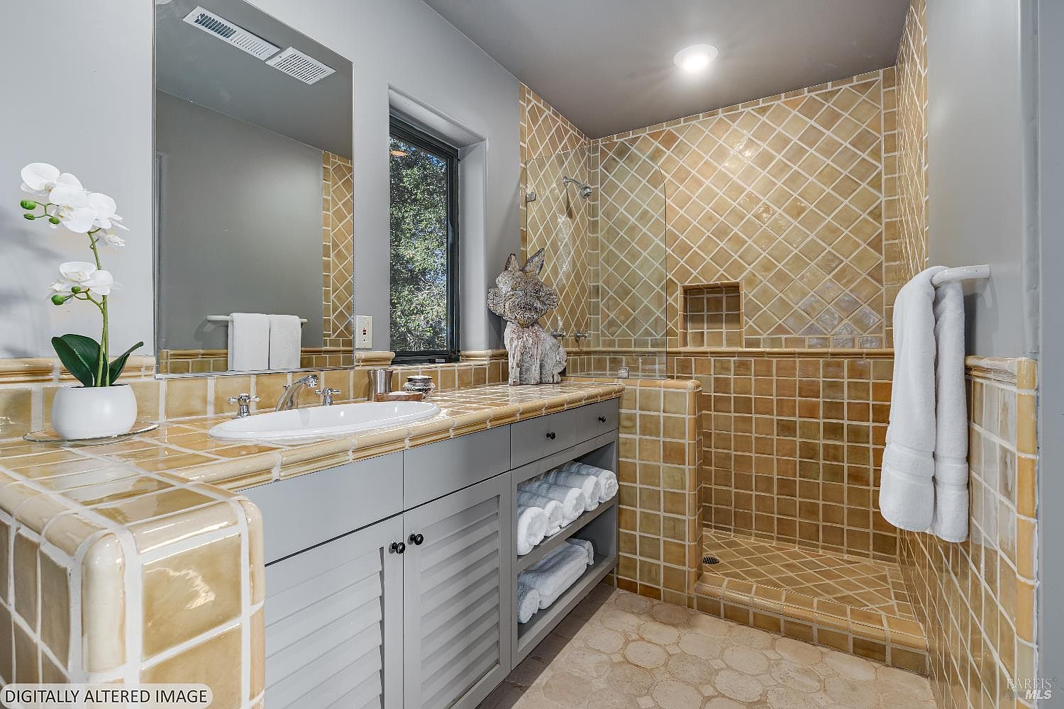 This bathroom features a unique, warm-toned aesthetic with tan, square-tiled countertops and a matching walk-in shower enclosure. The vanity is painted in a soft grey, complemented by open shelving filled with neatly stacked white towels and a large mirror above the sink. The space is well-lit by a recessed ceiling light and natural light streaming through a window, creating a cozy and inviting atmosphere.