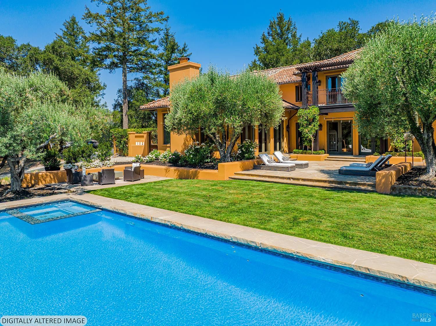 This stunning Mediterranean-style estate features a vibrant blue swimming pool and spa in the foreground, leading to a lush green lawn and a spacious stone patio. The rear of the home showcases warm, ochre-colored stucco walls, a terracotta tile roof, and mature olive trees that create a serene, resort-like atmosphere. The perspective captures the seamless integration of indoor-outdoor living, highlighting the property's luxurious and inviting backyard retreat.