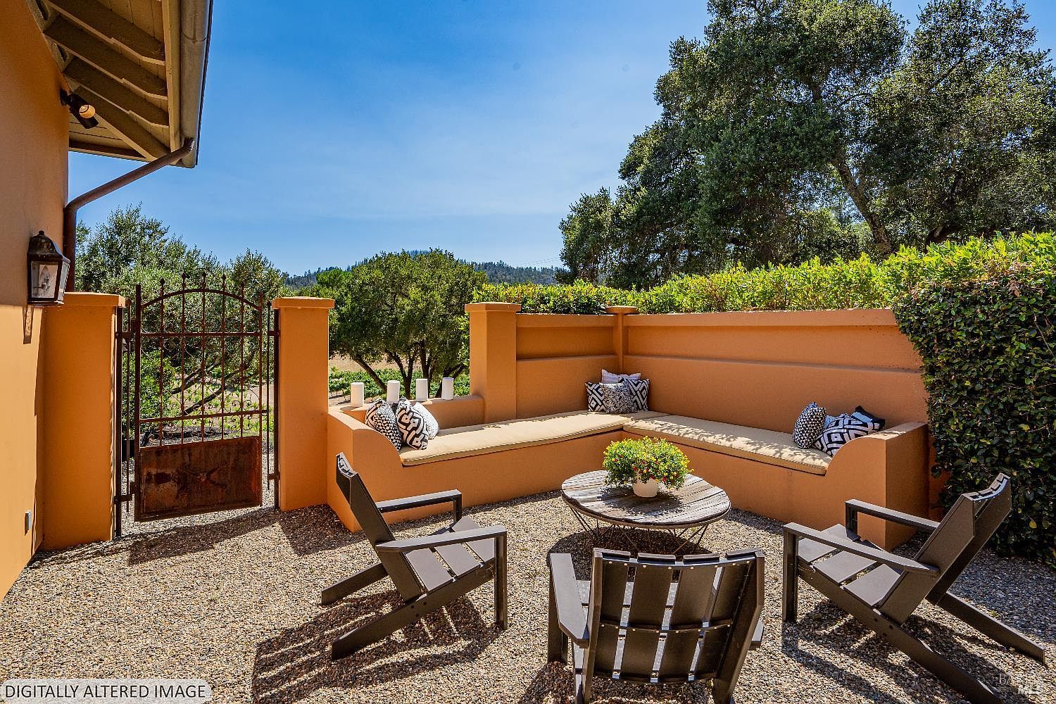 This inviting outdoor patio features a built-in L-shaped stucco bench with plush patterned cushions, centered around a rustic wooden coffee table. The space is furnished with three modern dark-toned chairs and is set on a gravel surface, framed by a wrought-iron gate and lush greenery. The warm, sun-drenched atmosphere creates a serene and private retreat perfect for relaxation or entertaining.