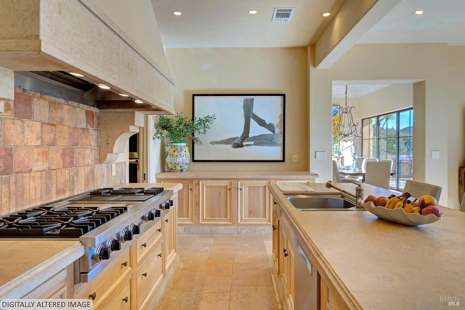 This spacious, high-end kitchen features a large central island with a stone countertop and a built-in sink, complemented by light wood cabinetry and a professional-grade range. A decorative tile backsplash adds texture behind the stove, while a large piece of abstract art hangs above a buffet area, creating a sophisticated focal point. The perspective is a wide-angle shot from the kitchen island looking toward the dining area, showcasing an open-concept layout with warm, inviting lighting.