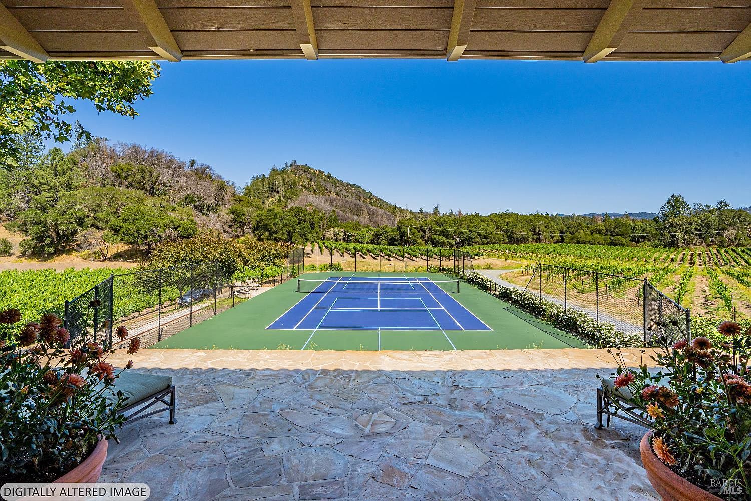This image captures a scenic view from a stone-paved patio overlooking a private tennis court nestled within a lush vineyard. The perspective is framed by an overhead wooden structure, creating a shaded foreground that leads the eye toward the vibrant blue and green court and the rolling hills in the distance. The setting offers a serene and luxurious outdoor living experience, perfect for recreation and enjoying the natural landscape.