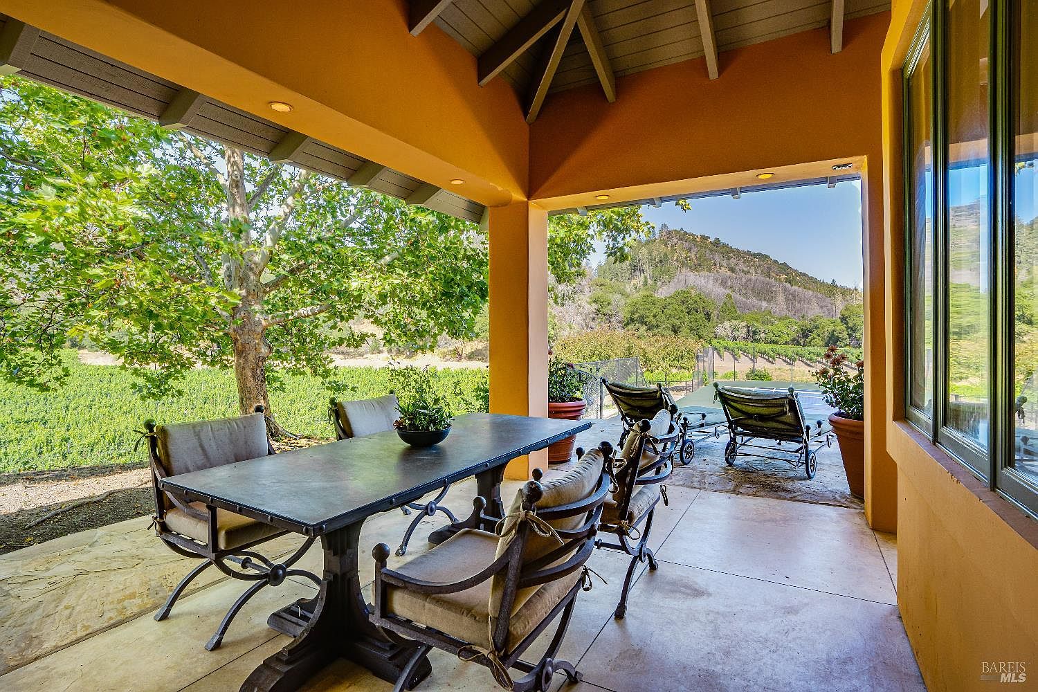 This covered outdoor patio offers a serene and sophisticated dining space, featuring a large metal table with comfortable cushioned chairs. The open-air design seamlessly connects the living area to a picturesque landscape of vineyards and rolling hills, creating an ideal setting for al fresco entertaining. The space is further enhanced by lounge chairs on the adjacent terrace, providing a perfect vantage point to enjoy the tranquil, scenic surroundings.