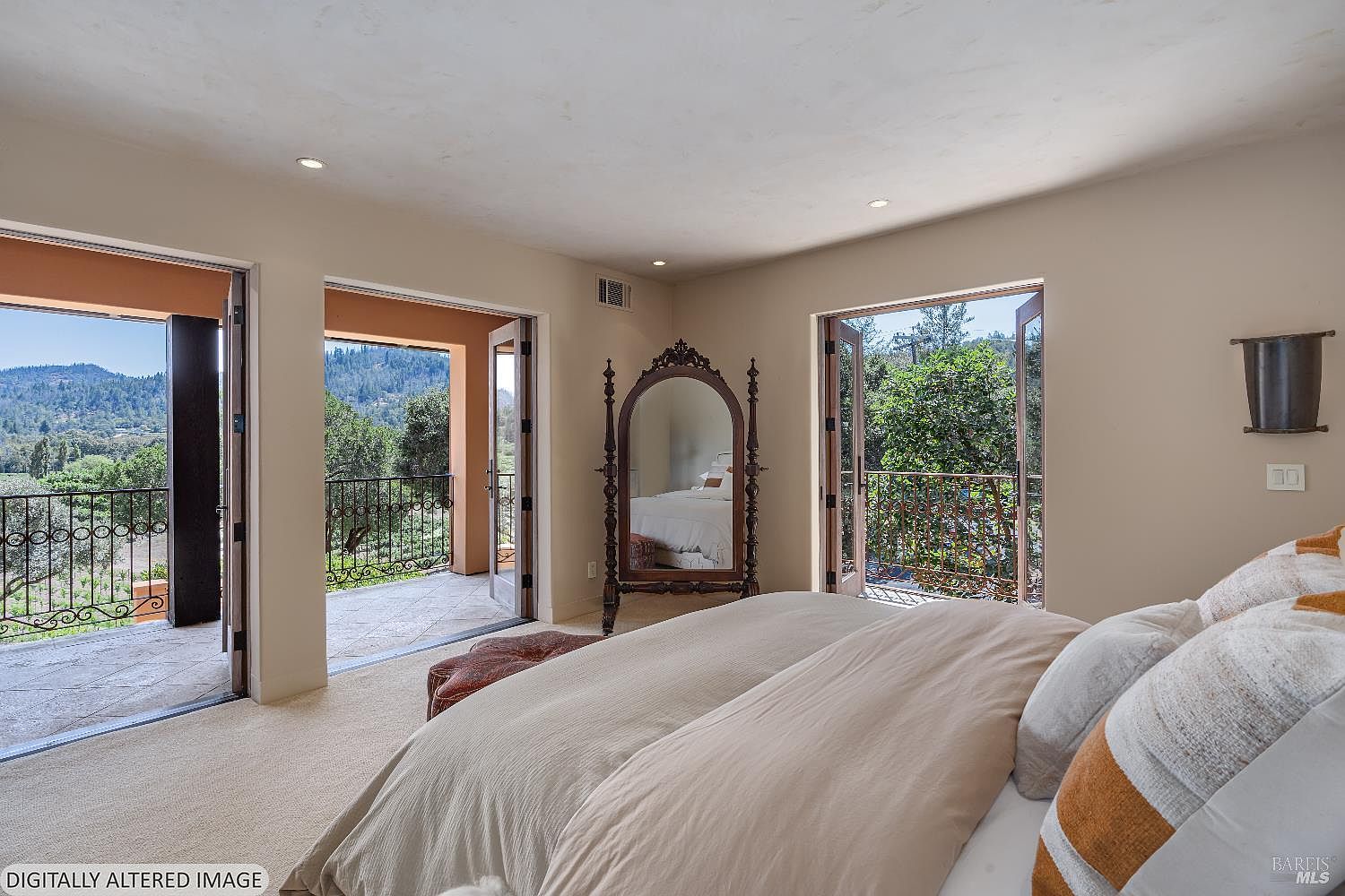 This primary bedroom features a neutral color palette and a plush bed with textured pillows, creating a serene and inviting atmosphere. The room is highlighted by two sets of French doors that open onto a private balcony, offering expansive views of the surrounding hills and landscape. A decorative floor mirror adds a touch of classic elegance to the space, which is bathed in natural light.