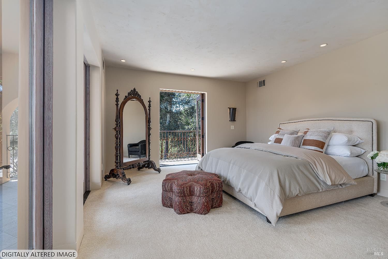 This primary bedroom features a neutral color palette with a plush, upholstered bed as the focal point, complemented by a decorative patterned ottoman at its foot. A large, ornate floor mirror stands to the left, while an open doorway leads to a private balcony, inviting natural light into the space. The room exudes a serene and sophisticated atmosphere, perfect for a relaxing retreat.