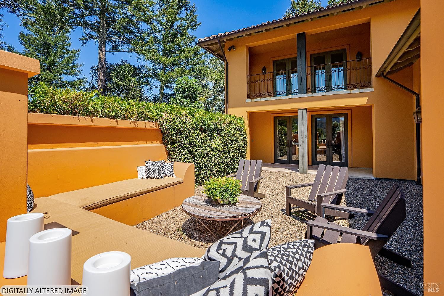 This inviting outdoor patio space features a built-in concrete bench with plush cushions, a rustic circular wooden coffee table, and several dark wood Adirondack chairs arranged on a gravel surface. The area is framed by a tall, manicured hedge and overlooks the rear facade of a two-story Mediterranean-style home with a balcony and French doors. The warm, sun-drenched atmosphere creates a serene and private retreat perfect for relaxation or entertaining.