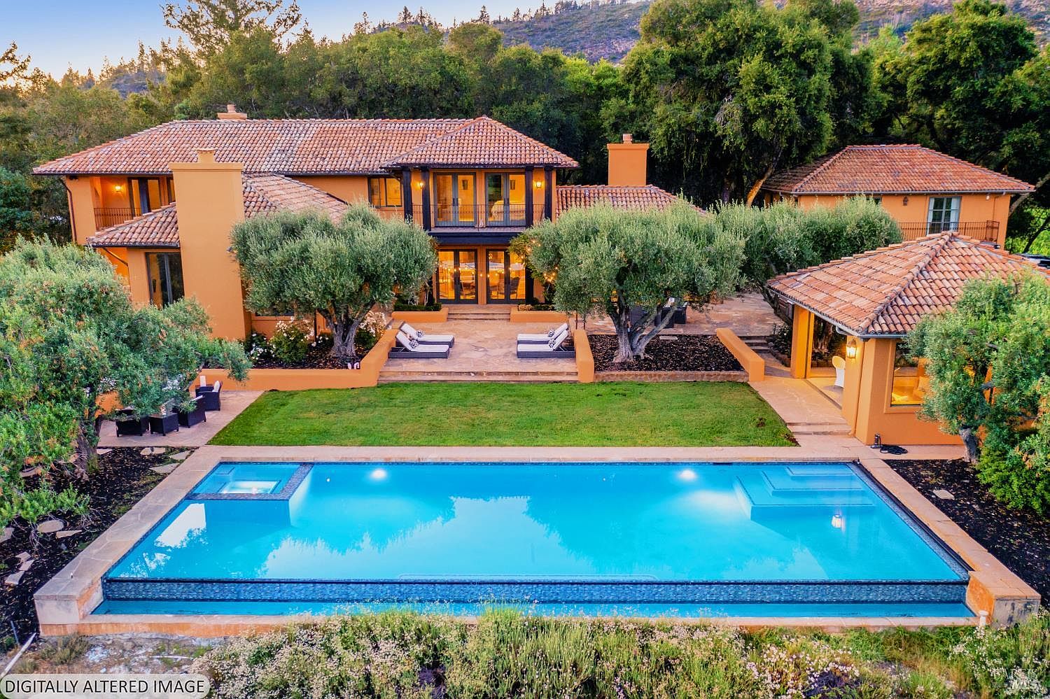 This stunning Mediterranean-style estate is captured from an elevated rear perspective, showcasing a sprawling backyard oasis. The property features a large, rectangular infinity-edge swimming pool with an integrated spa, a manicured lawn, and mature olive trees framing the home's warm, terracotta-toned facade. The architecture highlights classic tile roofing, multiple balconies, and a detached structure, creating a luxurious and serene outdoor living environment.