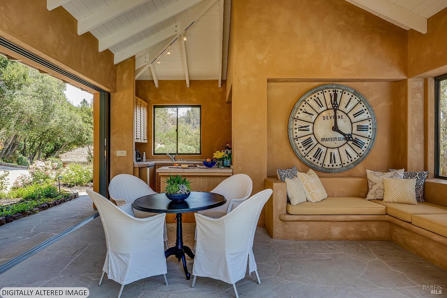 This inviting covered patio features a rustic, Mediterranean-inspired design with warm, earth-toned stucco walls and a stone floor. The space is furnished with a round dining table and chairs, a built-in bench with decorative pillows, and a large, statement wall clock, all opening seamlessly to the outdoors through a wide sliding glass door. The open-air layout creates a perfect transition between the indoor living area and the lush, sunlit garden beyond.