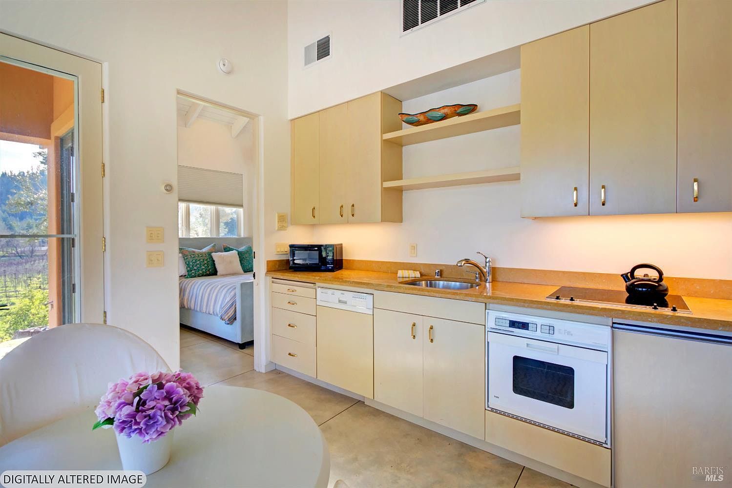 This bright and compact kitchen features light-toned cabinetry, a stone countertop, and integrated appliances, including a white oven and dishwasher. A small dining area with a floral centerpiece sits in the foreground, while an open doorway provides a clear view into an adjacent bedroom. The space is well-lit with under-cabinet lighting and offers a scenic view of the outdoors through a glass door.