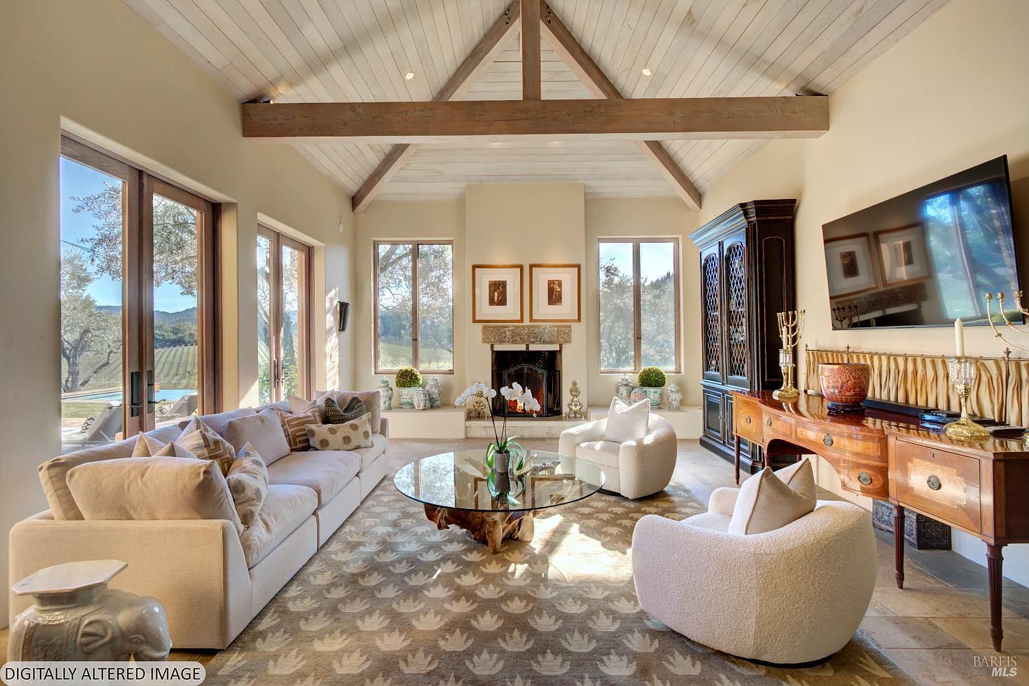 This elegant living room features a high, vaulted ceiling with exposed wooden beams and white shiplap, creating a bright and airy atmosphere. The space is anchored by a stone fireplace, a large glass-topped coffee table with a natural wood base, and plush, neutral-toned seating, all set against a backdrop of expansive windows that offer views of the surrounding vineyard. The room exudes a sophisticated, rustic-chic aesthetic, perfectly blending luxury with the natural beauty of the outdoors.