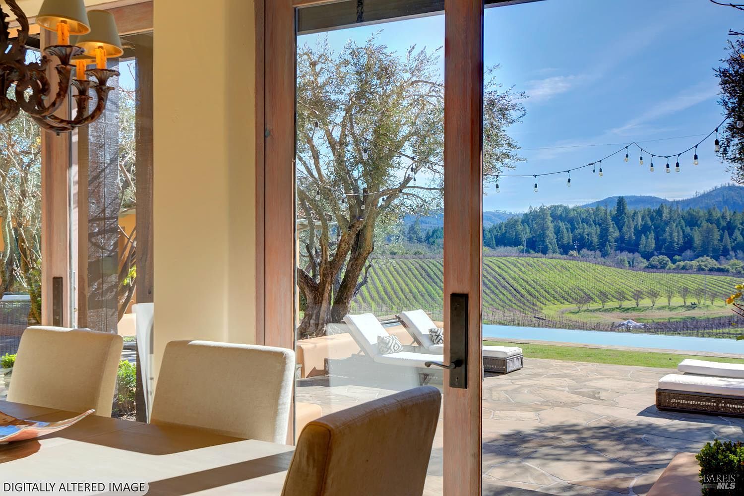 This interior view captures a sophisticated dining area that seamlessly transitions to an outdoor patio through large glass sliding doors. The foreground features elegant dining chairs and a chandelier, while the background showcases a stunning vineyard landscape, a swimming pool, and lounge chairs. The perspective emphasizes the indoor-outdoor lifestyle, highlighting the expansive views and natural light that define this luxury property.