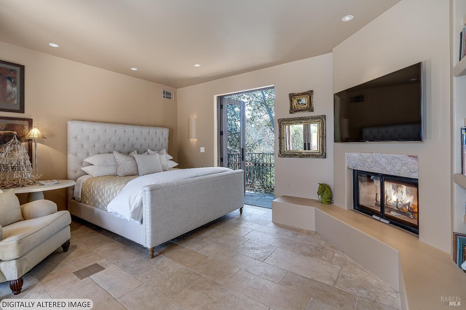 This elegant primary bedroom features a plush, tufted upholstered bed as the centerpiece, complemented by a cozy armchair and a warm, inviting fireplace. The room is bathed in natural light from the open French doors that lead to a private balcony, creating a seamless indoor-outdoor living experience. The neutral color palette, stone tile flooring, and tasteful decor contribute to a sophisticated and tranquil atmosphere.