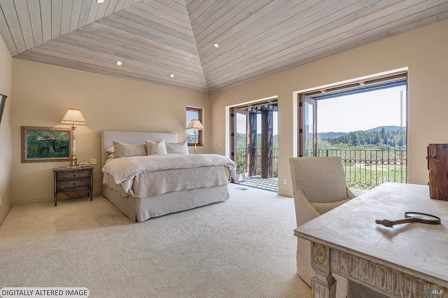 This spacious primary bedroom features a vaulted, light-wood paneled ceiling and a neutral color palette that creates a serene, airy atmosphere. The room is furnished with a plush king-sized bed, a classic wooden nightstand with a lamp, and a desk area, all complemented by large glass doors that open onto a balcony with scenic vineyard views. The perspective is from the corner of the room, capturing both the sleeping area and the workspace, emphasizing the bright, natural light and elegant design.