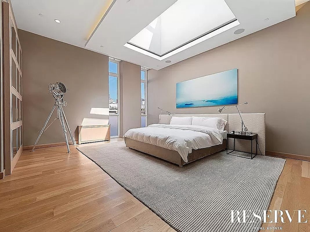 This spacious primary bedroom features light-toned walls, warm wood flooring, and a large area rug centered under a platform bed. A prominent skylight above the bed floods the room with natural light, complemented by tall windows along the side wall. A unique tripod-style floor lamp adds a modern, artistic touch to the minimalist aesthetic of the room.