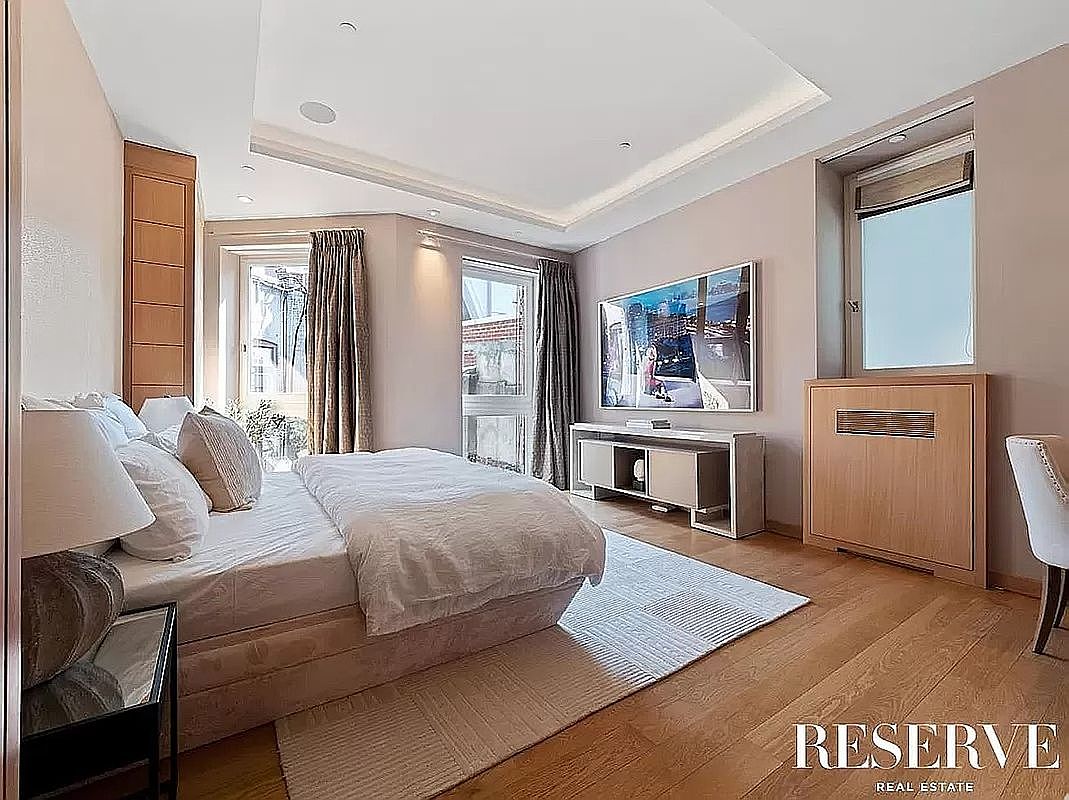 This primary bedroom features a neutral color palette with soft beige walls and light wood flooring, creating a serene and sophisticated atmosphere. The room is furnished with a plush bed, a sleek media console beneath a large framed artwork, and a built-in wooden storage unit. Large windows with elegant curtains allow natural light to fill the space, highlighting the clean lines and modern aesthetic of the room.