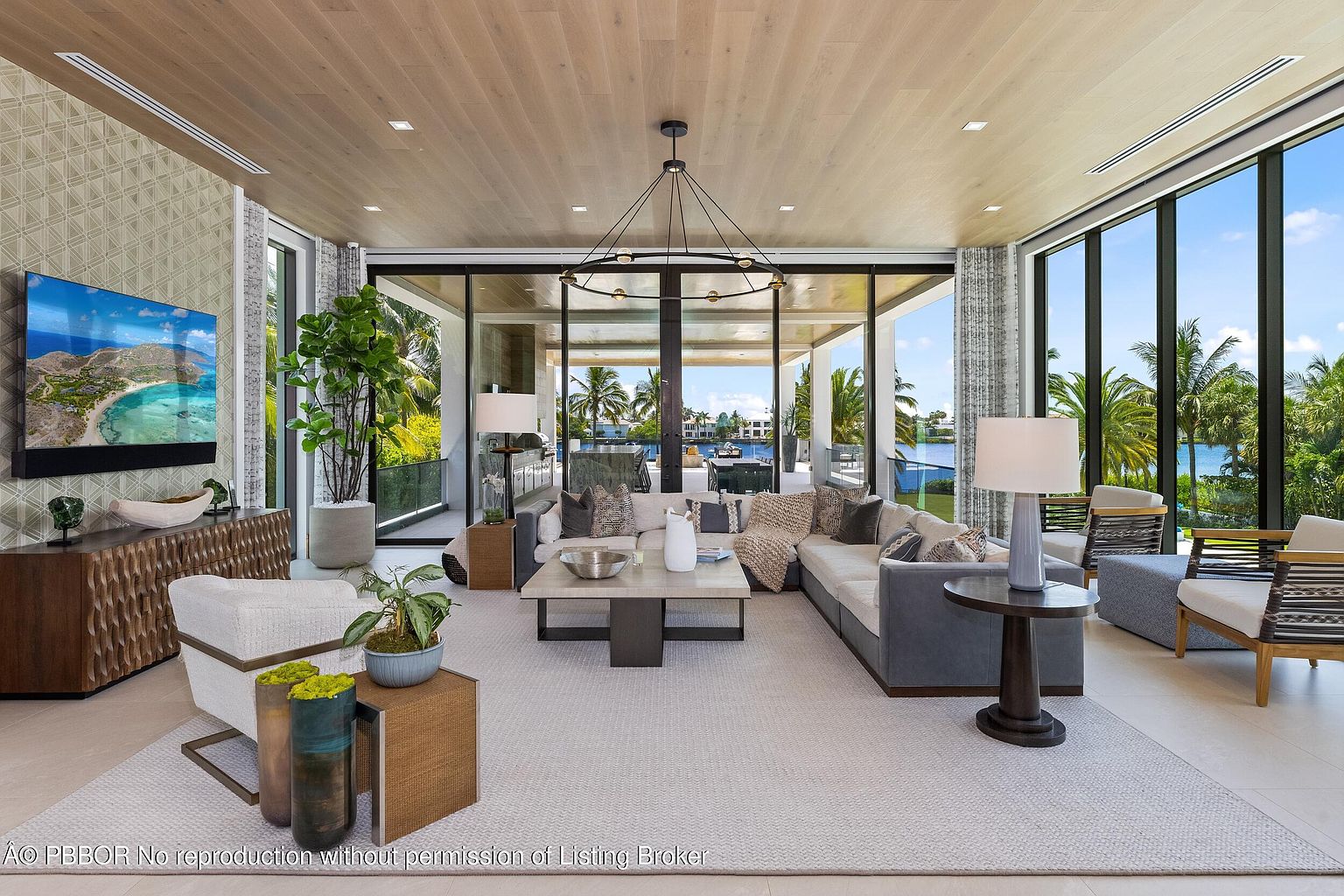 This is a luxurious living room featuring a large sectional sofa, a modern chandelier, and floor-to-ceiling windows offering stunning waterfront views. The room is decorated in neutral tones with wood accents, creating a bright and airy atmosphere. The open floor plan and high ceilings enhance the sense of spaciousness, making it an ideal space for relaxation and entertaining.
