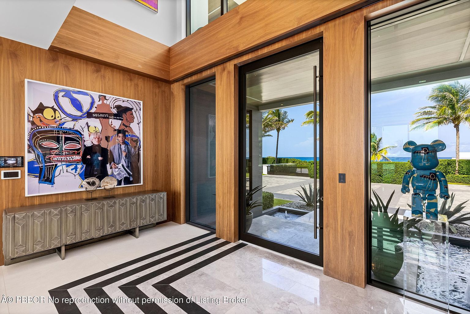 This interior shot showcases a modern hallway with striking architectural details. The space features wood-paneled walls, a bold black and white geometric floor pattern, and a contemporary art piece above a stylish console table. Large glass doors offer a glimpse of the exterior, creating a seamless transition between indoor and outdoor spaces.