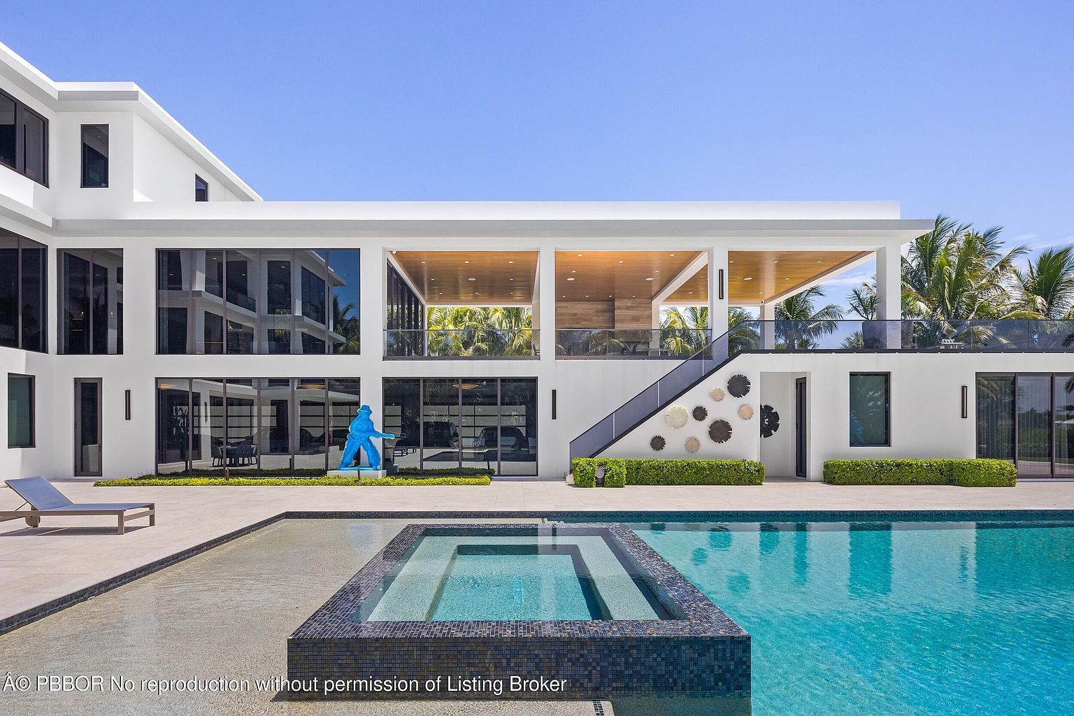 This image showcases the rear exterior of a modern luxury home, emphasizing the pool and spa area. The pool features a raised spa with dark blue tiling, and the home's architecture includes large windows, a covered outdoor living space on the second floor, and a contemporary design with clean lines. A blue sculpture adds a pop of color to the meticulously landscaped yard.