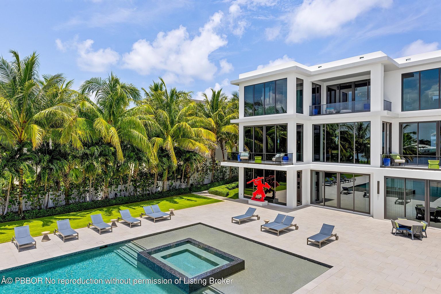 This image showcases the rear exterior of a modern luxury home, emphasizing the pool and spa area. The property features a sleek, white facade with expansive windows, a patio with lounge chairs, and lush tropical landscaping. A striking red sculpture adds a pop of color and artistic flair to the outdoor space.