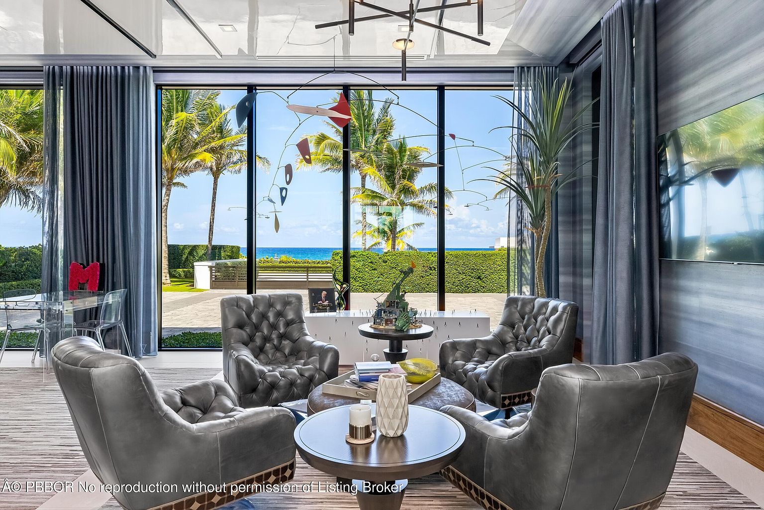 This is an interior shot of a luxurious living room featuring four tufted leather armchairs arranged around a dark wood coffee table and ottoman. A large window provides a stunning view of palm trees, the ocean, and manicured landscaping, while a modern mobile hangs in front of the window. The room is decorated with high-end finishes and stylish accessories, creating an inviting and sophisticated atmosphere.