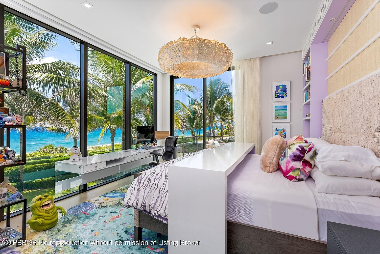 This is a bright and airy bedroom featuring a modern design with a playful touch. The room boasts large windows offering stunning ocean views framed by palm trees, a unique overhead light fixture, and a built-in desk area. The decor includes colorful artwork and whimsical accents, creating a cheerful and inviting space.