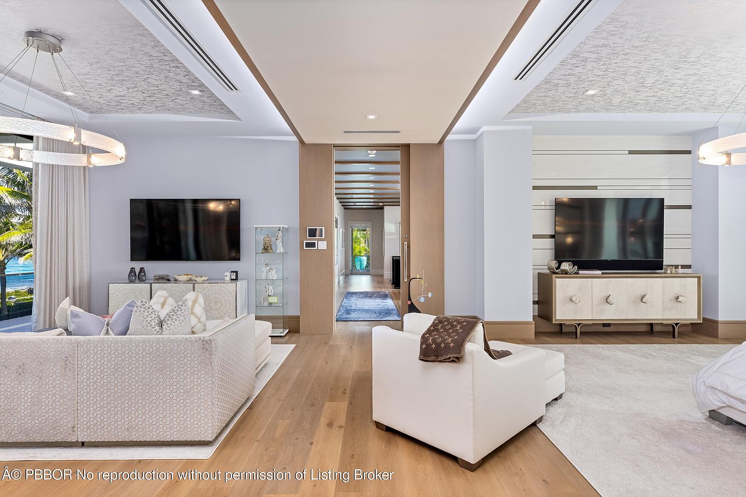 This is a luxurious living room featuring a modern design with high-end finishes. The space is divided into two distinct areas, each with its own television and seating arrangement. The room boasts hardwood floors, custom ceiling details, and a neutral color palette, creating an elegant and inviting atmosphere.