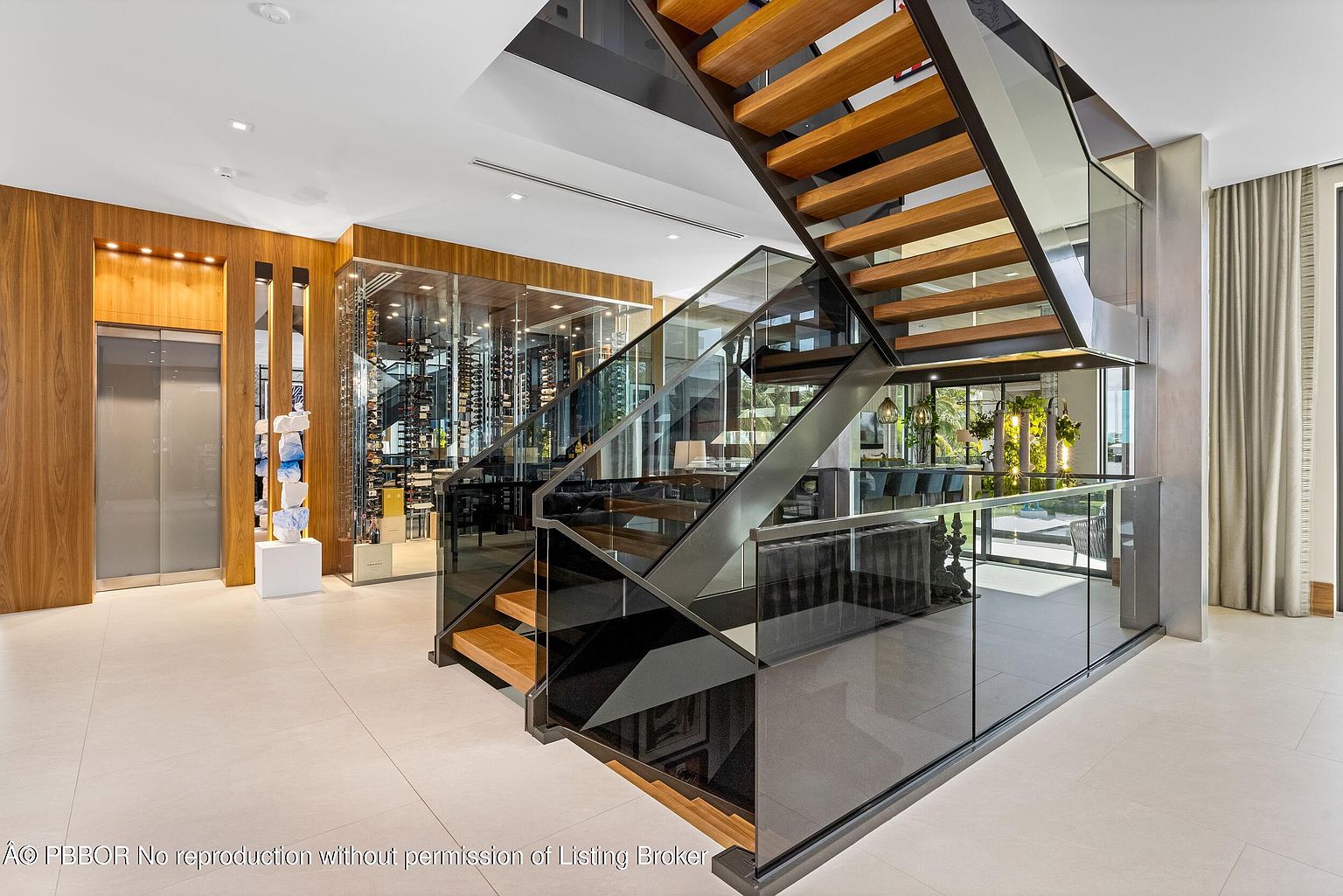 This interior shot showcases a modern hallway featuring a striking staircase with wooden steps and glass railings. A glass-enclosed wine cellar is visible in the background, adding a luxurious touch. The space is well-lit with a clean, contemporary design, emphasizing openness and sophistication.
