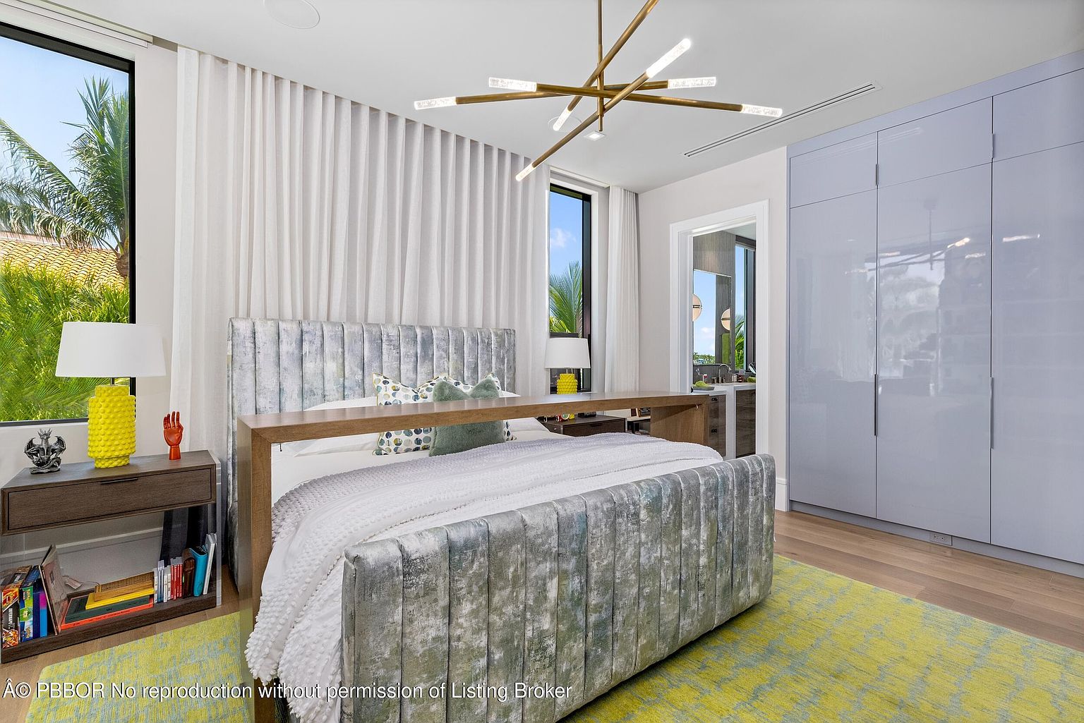 This is a primary bedroom featuring a unique bed with a wooden shelf-like headboard and footboard, complemented by a modern chandelier. The room includes a large window with sheer curtains, a bedside table with a yellow pineapple lamp, and built-in gray wardrobes. The overall style is contemporary and luxurious, with a bright and airy feel.