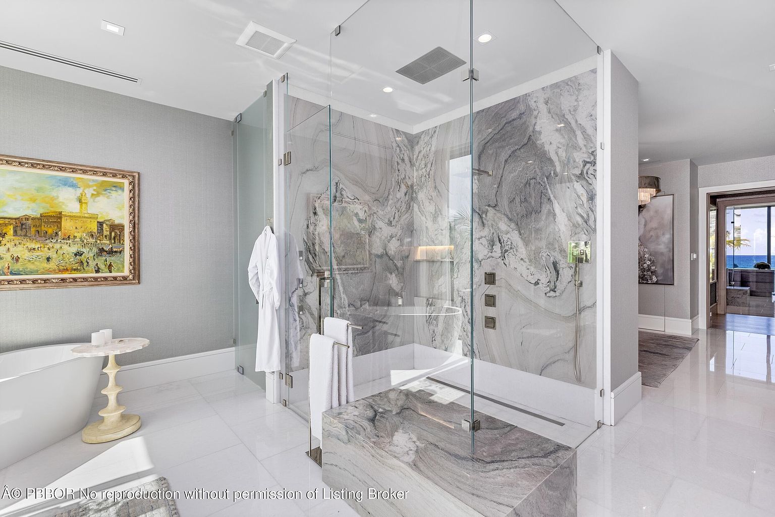 This is a luxurious primary bathroom featuring a glass-enclosed shower with marble walls and modern fixtures. A freestanding bathtub sits adjacent to a decorative table with a painting on the wall, adding an artistic touch. The room is bright and spacious, with glossy white tile flooring that enhances the clean and elegant aesthetic.