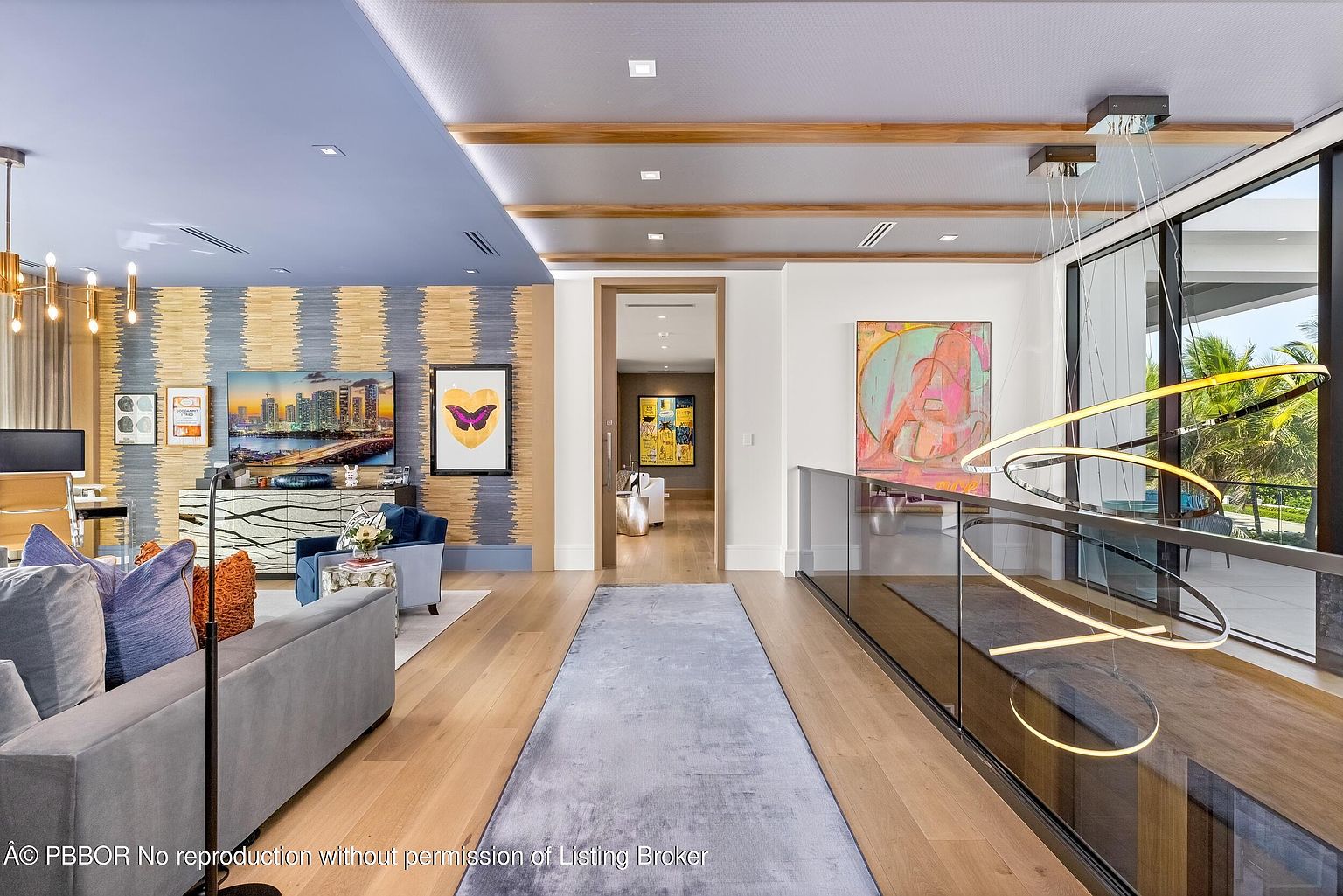 This interior shot showcases a luxurious hallway connecting living spaces. The hallway features hardwood flooring, a long area rug, and modern lighting fixtures. A glass railing provides a view of the lower level, while artwork adorns the walls, creating an elegant and inviting atmosphere.