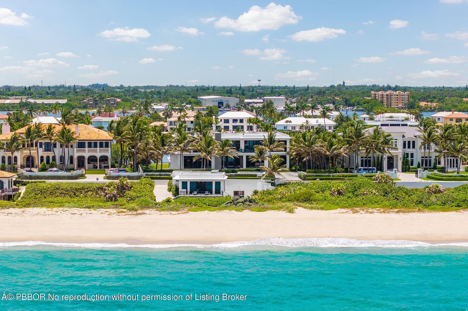 This aerial view showcases a luxurious beachfront property with multiple buildings, lush landscaping, and direct access to a pristine sandy beach and turquoise ocean. The architectural style is modern and elegant, with palm trees adding a tropical touch. The image conveys a sense of exclusivity and high-end coastal living.