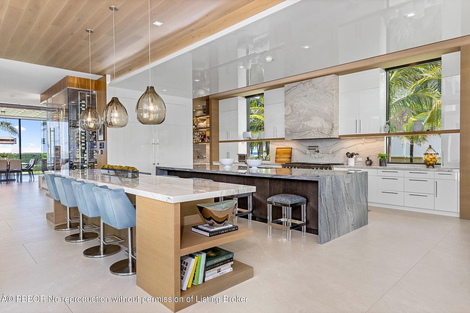 This is a bright and modern kitchen featuring a large island with seating, marble countertops, and sleek white cabinetry. The kitchen includes high-end appliances and a wine cellar visible in the background. The open layout and natural light create an inviting and luxurious atmosphere.