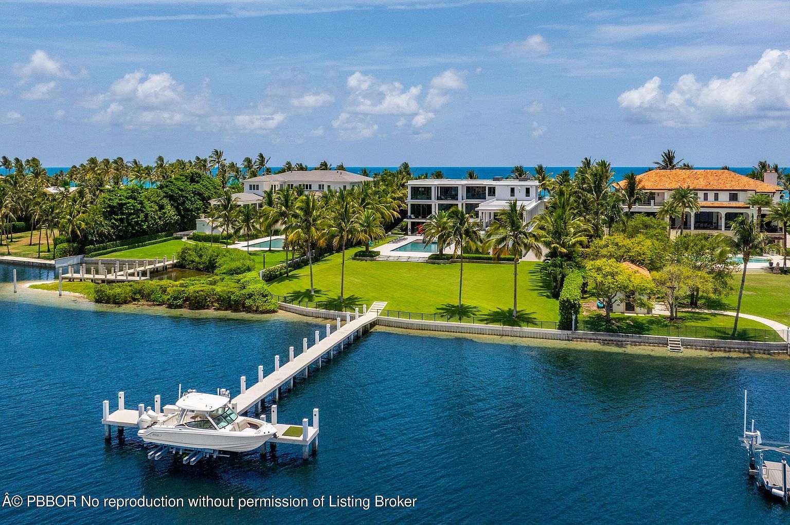 This aerial shot showcases a luxurious waterfront property featuring multiple residences with manicured lawns, palm trees, and private docks. A boat is moored at one of the docks, emphasizing the property's direct water access. The overall impression is one of opulence and serene coastal living.