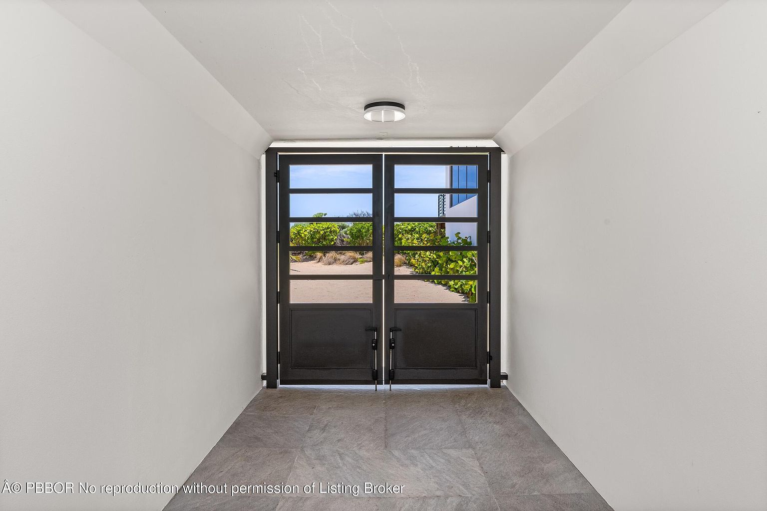 This interior shot showcases a hallway with a set of black metal and glass double doors at the end, providing a glimpse of the outdoors. The walls are painted in a neutral tone, and the floor is covered with large gray tiles. A modern ceiling light fixture illuminates the space, creating a clean and minimalist aesthetic.