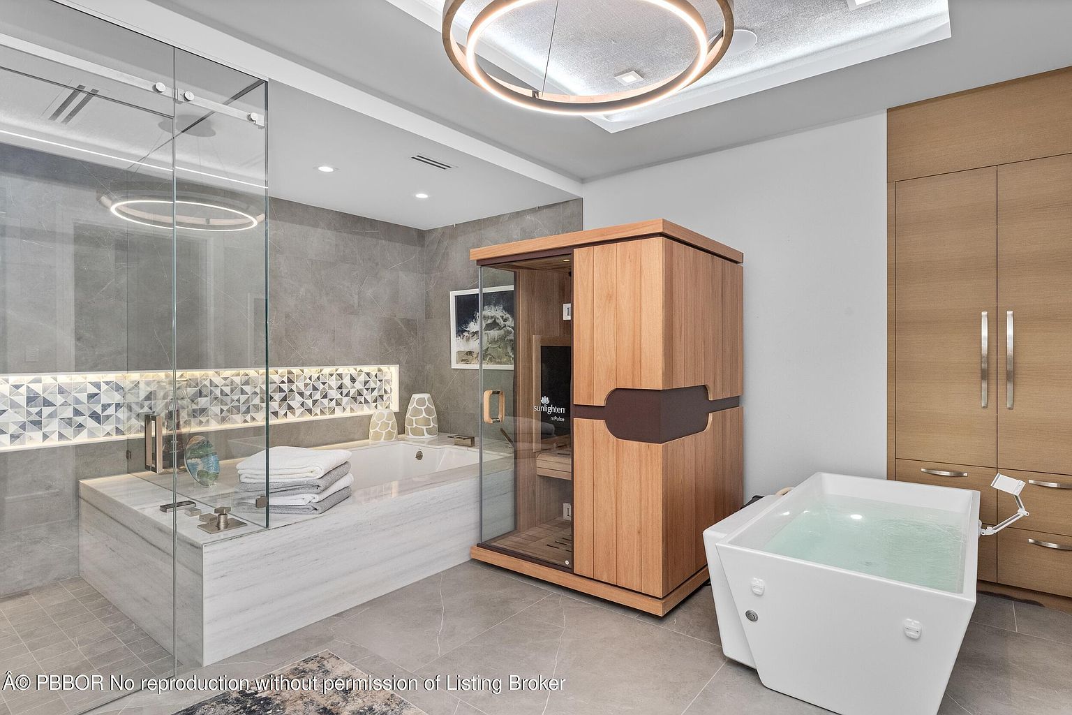 This is a luxurious primary bathroom featuring a modern design with a large soaking tub encased in marble, a glass-enclosed sauna, and a separate jacuzzi tub. The room is illuminated by a contemporary circular light fixture, and the neutral color palette creates a serene and spa-like atmosphere. The presence of high-end fixtures and finishes suggests a premium property.