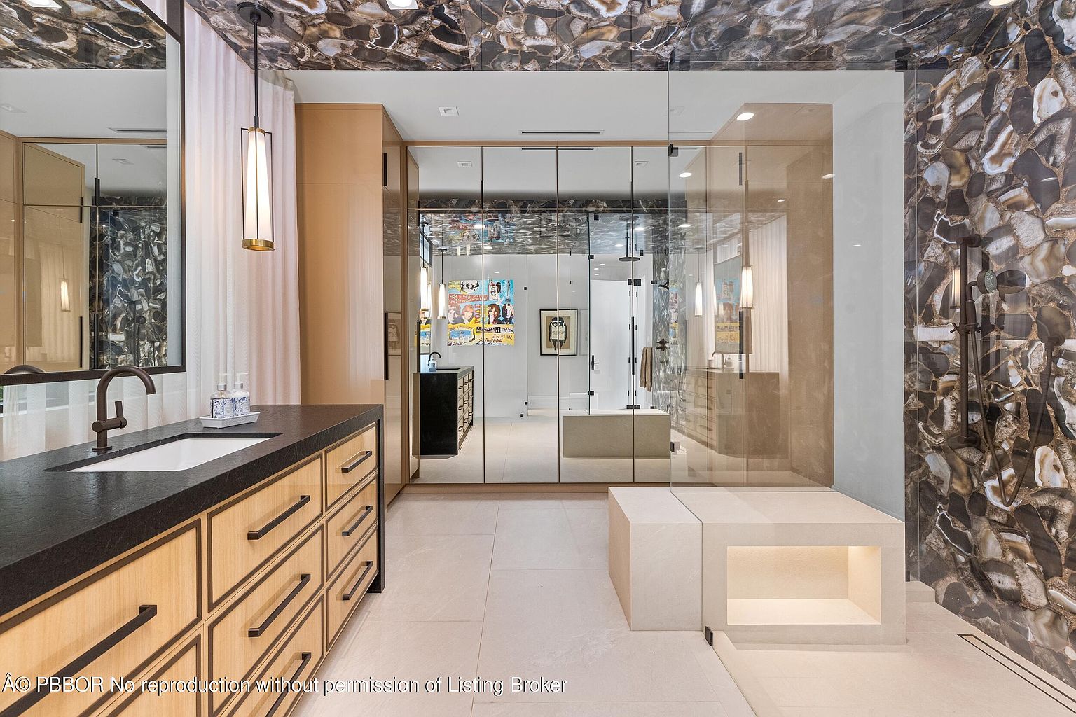 This is a luxurious primary bathroom featuring a unique stone ceiling and wall accent. The bathroom includes a vanity with a dark countertop and light wood drawers, a glass-enclosed shower area with a built-in bench, and reflective surfaces that enhance the sense of space. The overall design is modern and sophisticated, emphasizing high-end materials and finishes.