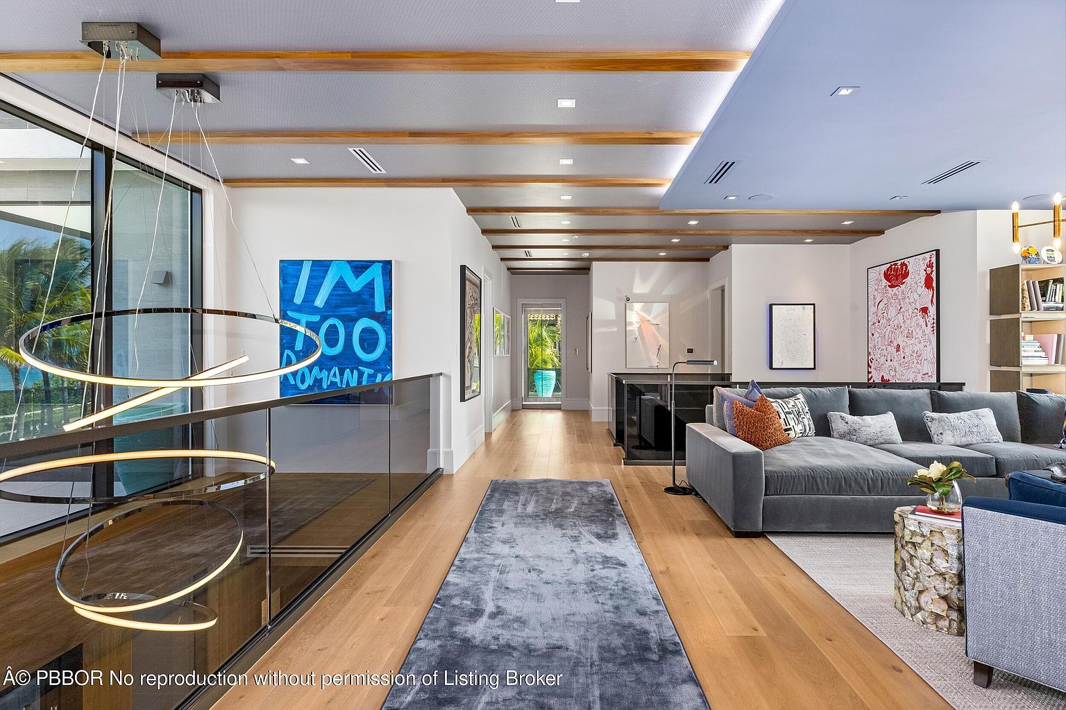 This interior shot showcases a modern hallway with wooden floors and a glass-railed staircase. The hallway features recessed lighting, wooden beam accents on the ceiling, and various pieces of contemporary art adorning the walls. A gray rug runs the length of the hallway, leading towards a living area with a gray sectional sofa.