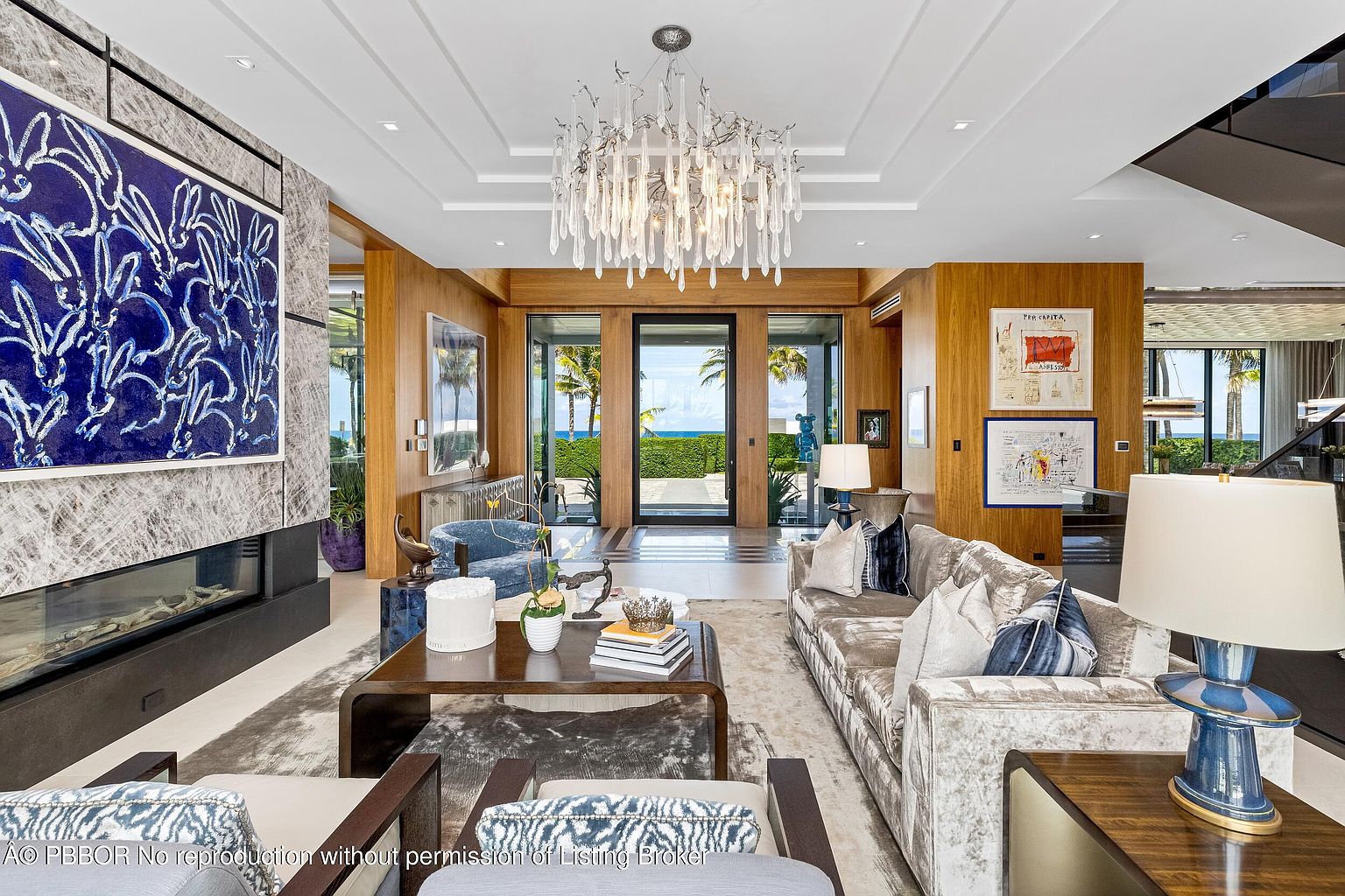 This is a luxurious living room featuring a modern chandelier, a large abstract painting above a fireplace, and comfortable seating. The room has wood paneling and large windows that offer a view of the ocean. The overall impression is elegant and sophisticated.